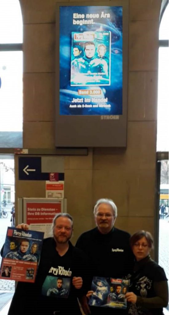 Thomas Seidel, Dieter Hofher und Michelle Stern posieren vor dem Werbebildschirm im Mainzer Hauptbahnhof. © Thomas Seidel